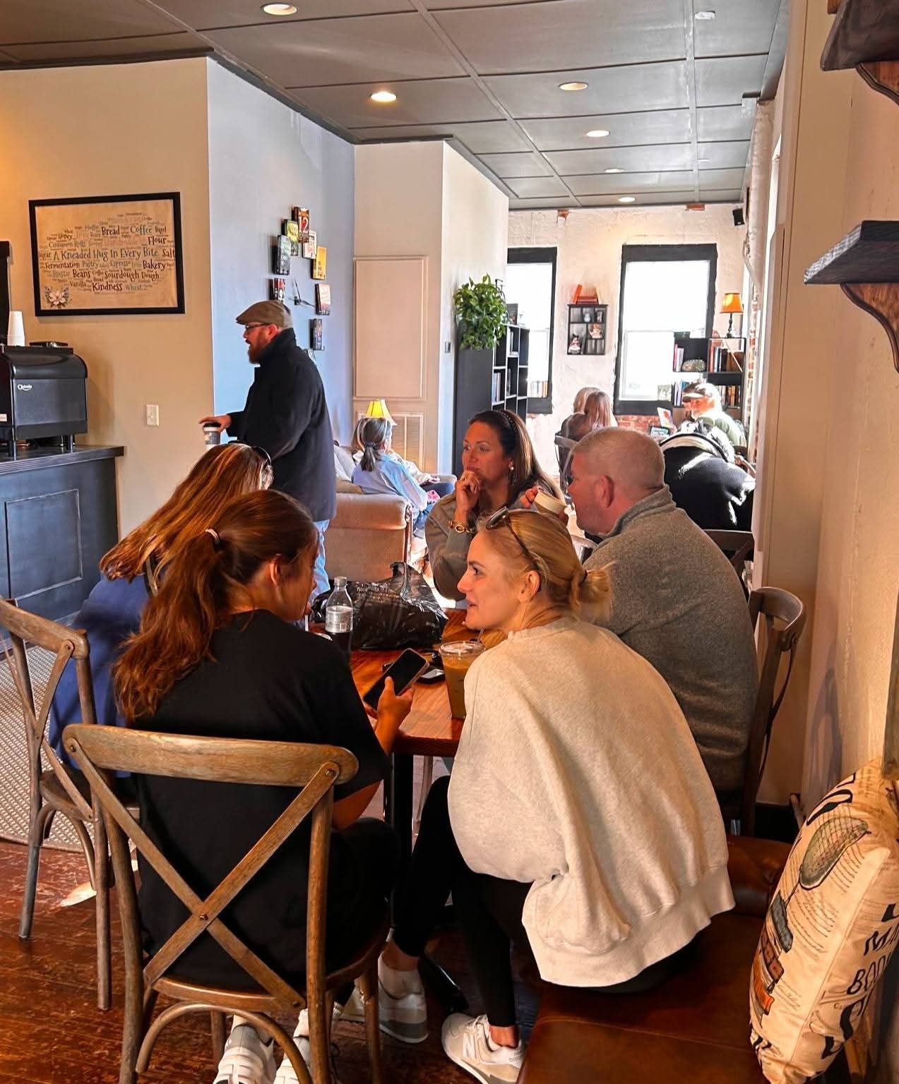People sit at tables inside a cafe, chatting and looking at phones in a cozy, softly lit room with a dark ceiling.