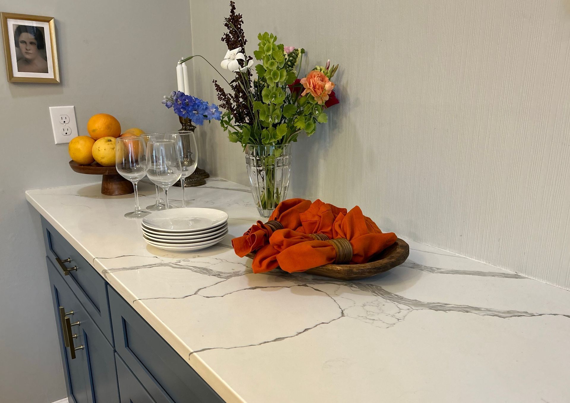 A kitchen counter with plates , wine glasses , and flowers on it.