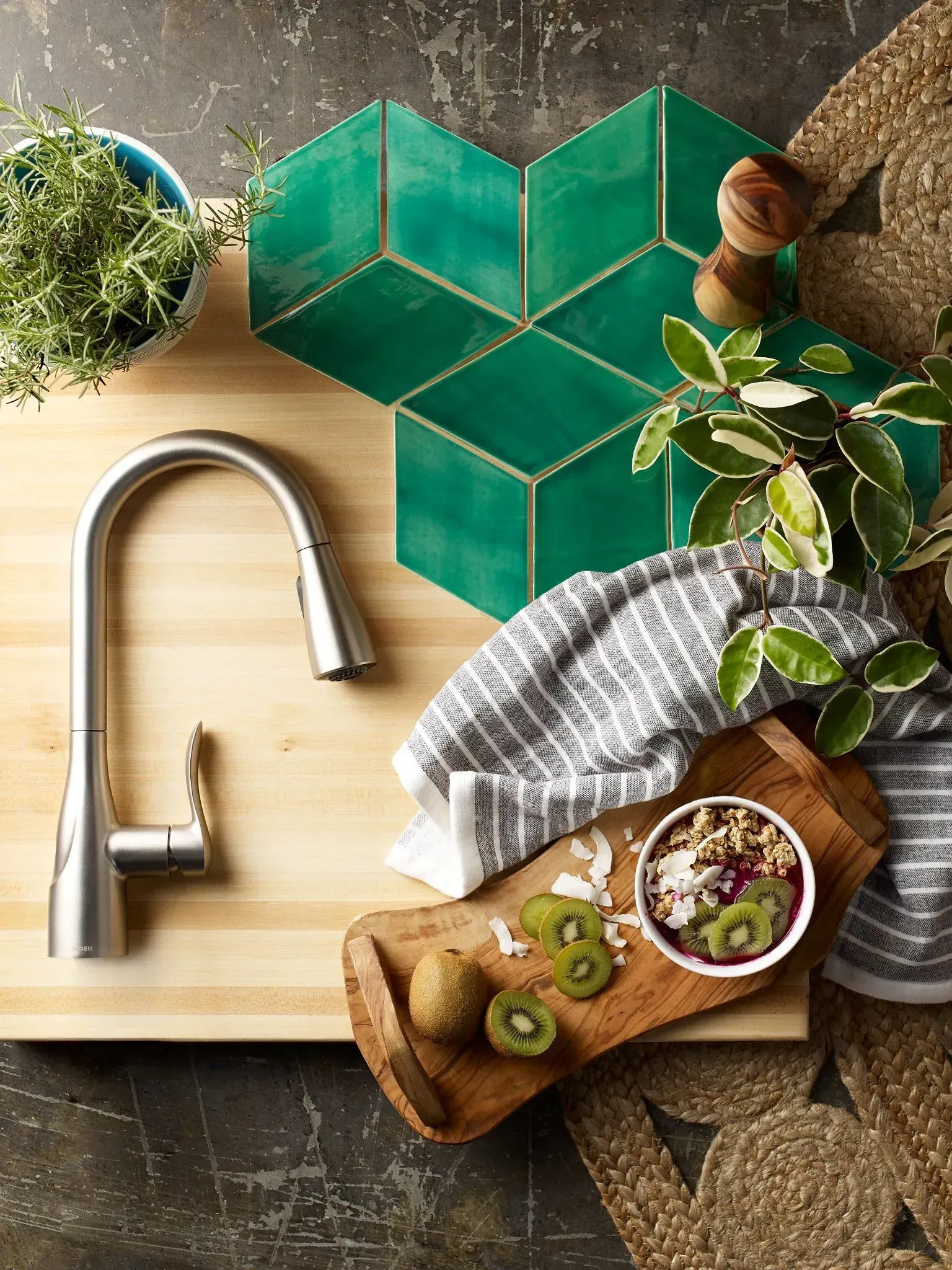 A kitchen counter with a faucet , cutting board , bowl of food and green tiles.