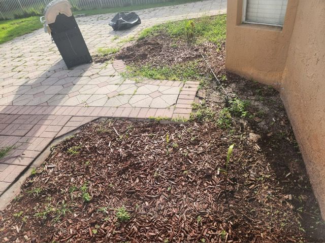 A trash can is sitting on the side of a house next to a brick walkway.