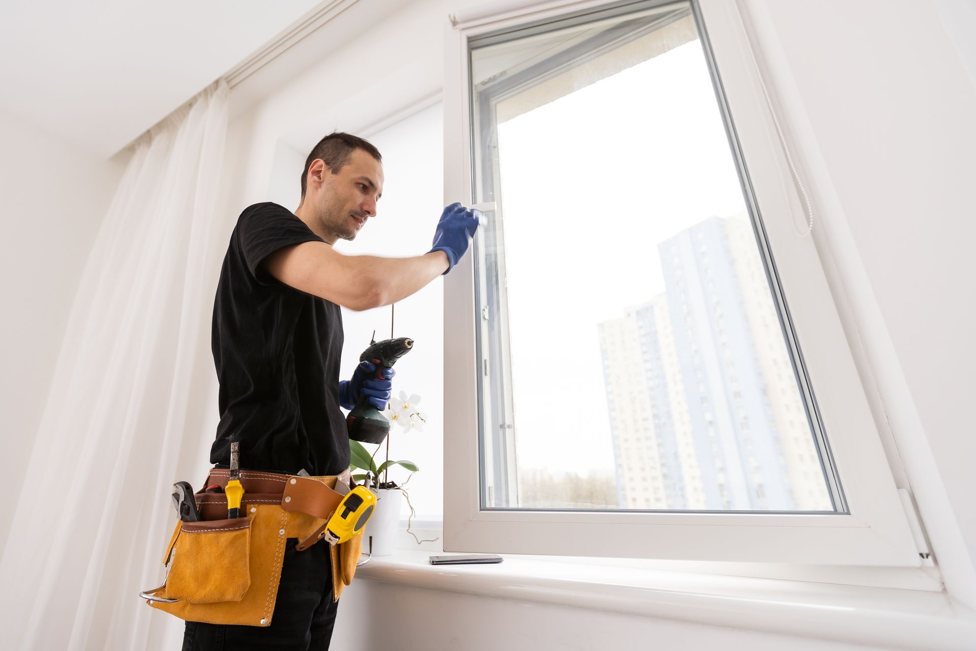 Man in work clothes repairing a window, using a drill, inside a white room, with city view outside.