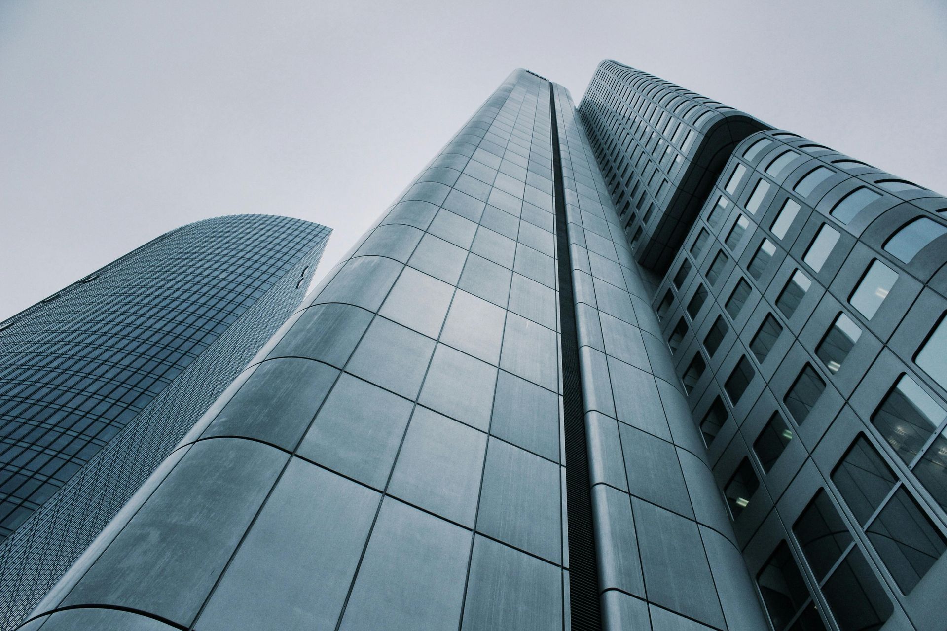 Low-angle view of three modern skyscrapers with metallic silver and blue panels against a cloudy sky.