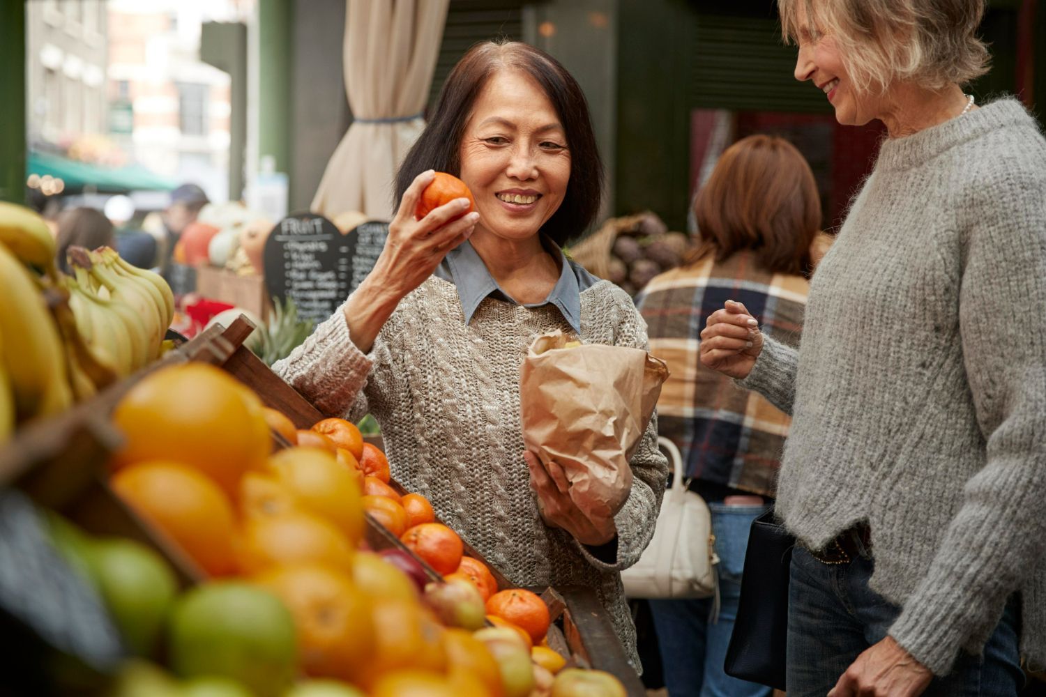 Woman at a market smiles, holding an orange, with another person and fruit visible.