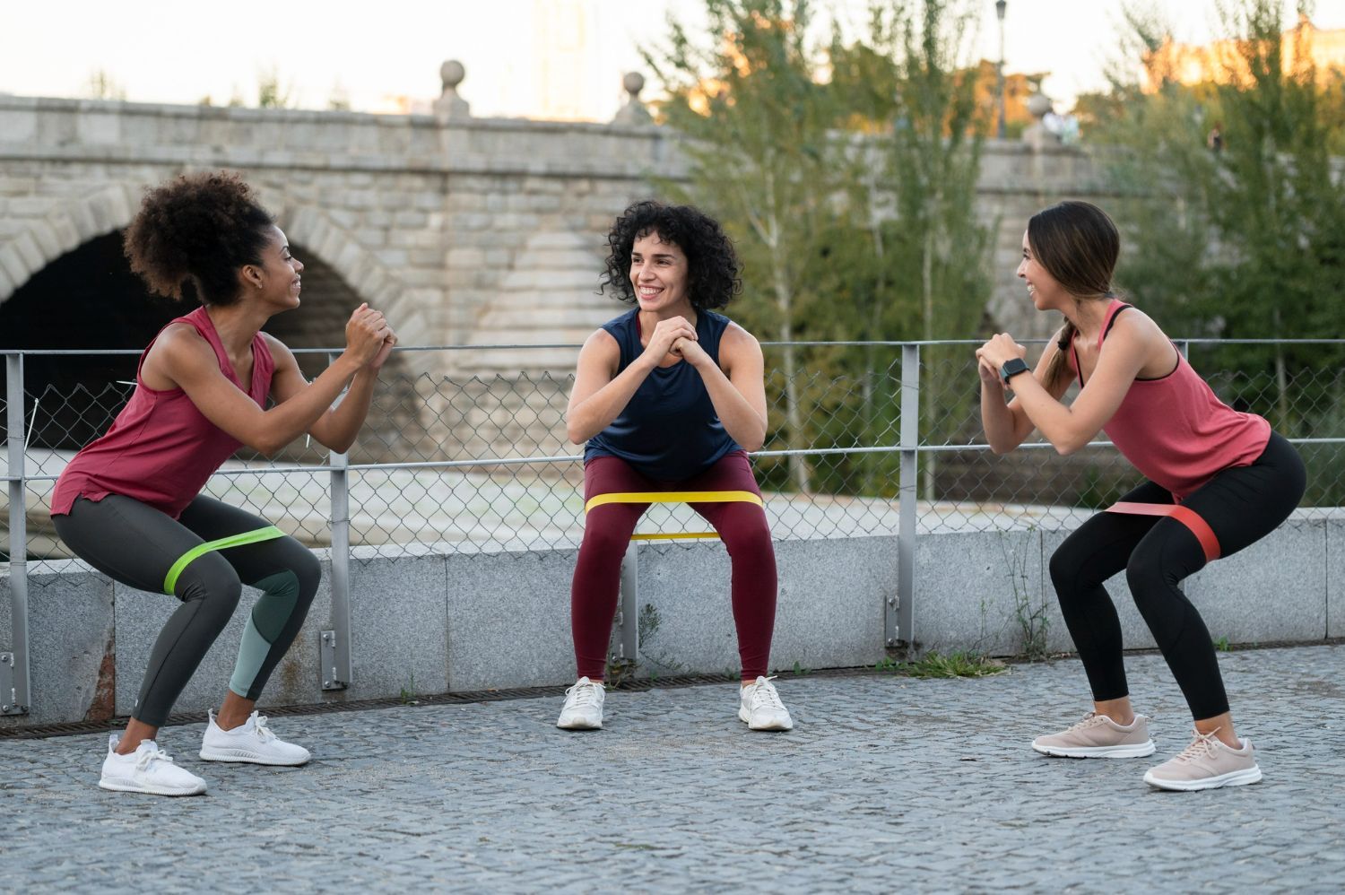 Three women doing squats with resistance bands outside.