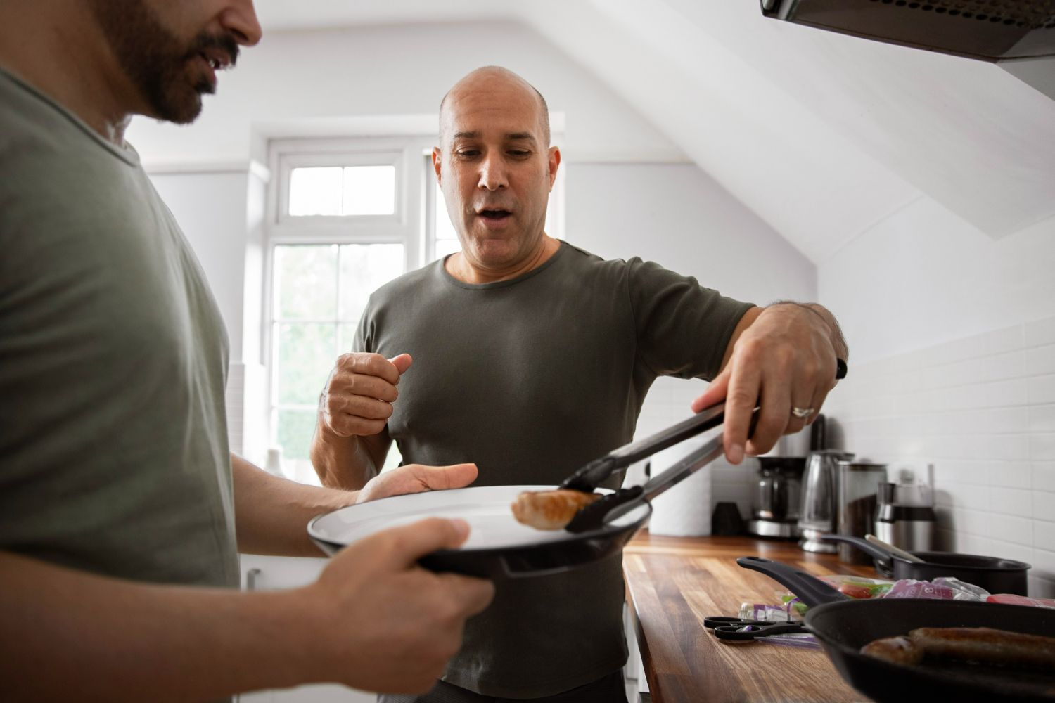 Two men cooking, one using tongs to place sausage on a plate held by the other in a kitchen.