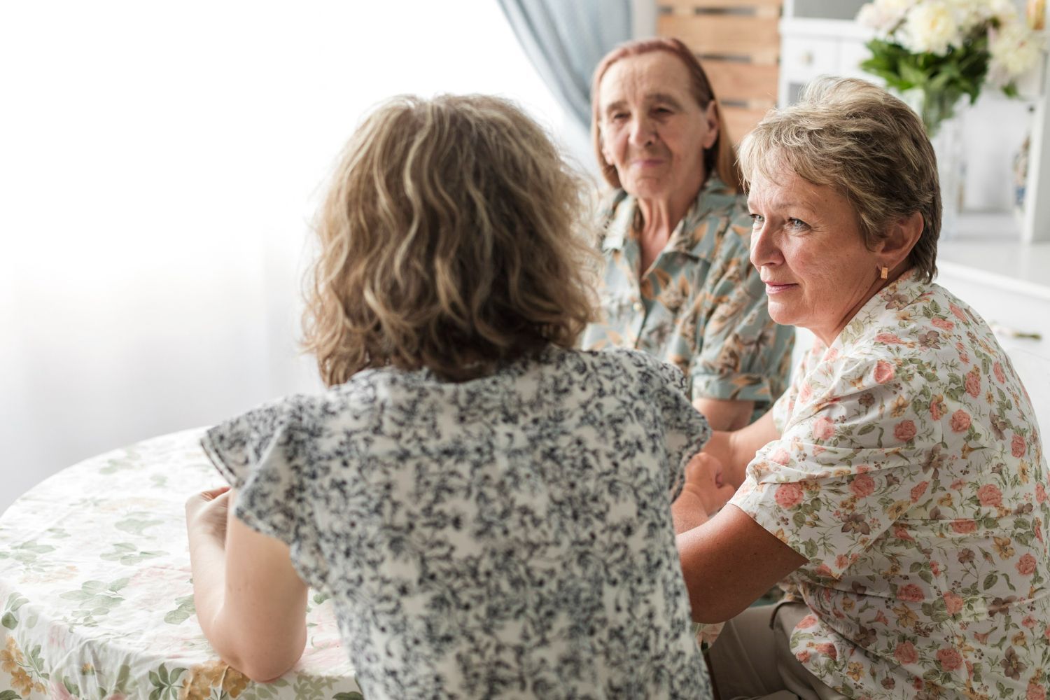Three women sit at a round table, talking. Floral tops, natural light, smiling, soft focus.
