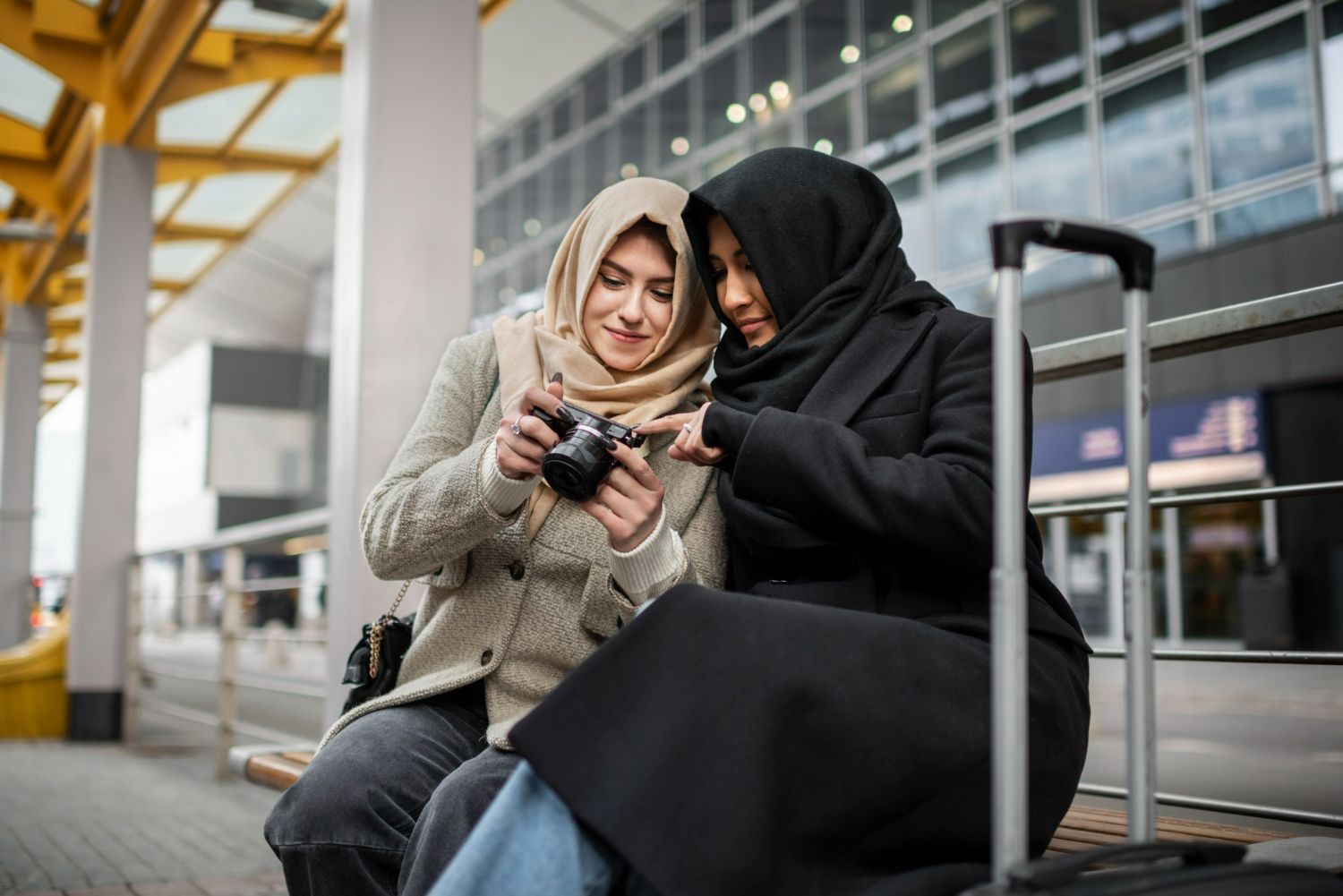 Two women in hijabs look at a camera, seated outdoors near luggage.