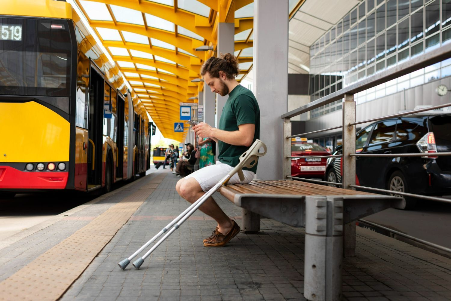 Man with crutches sits on bench at a bus stop, looking at phone. A bus and other vehicles are nearby.
