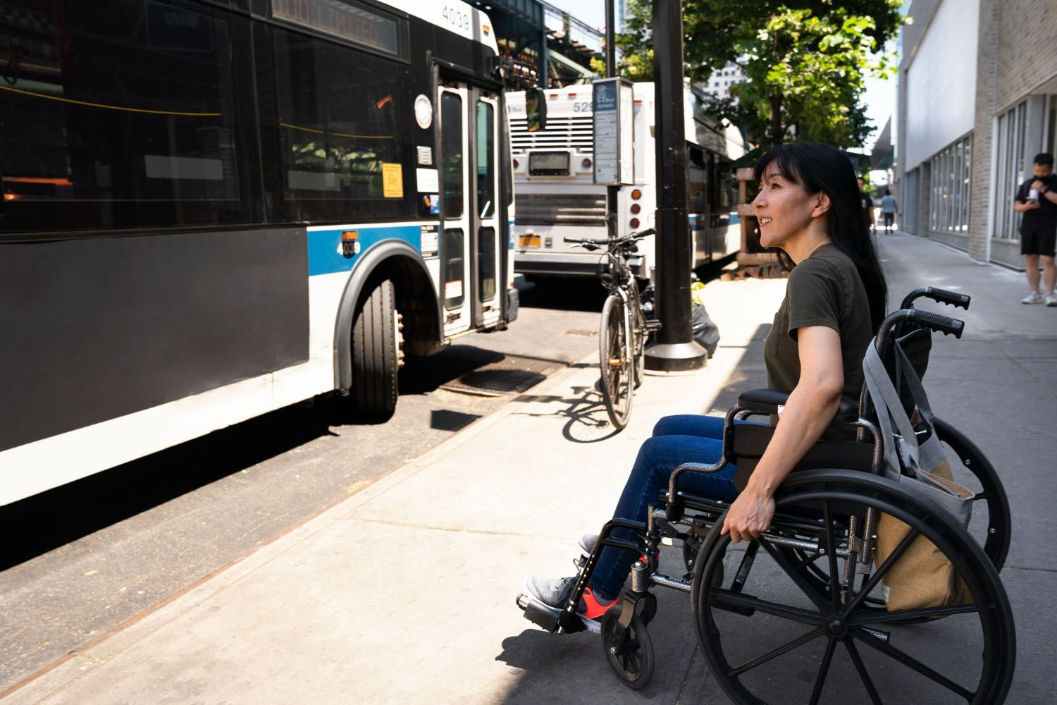 Woman in wheelchair waits for a city bus at a curb. Bright sunlight.