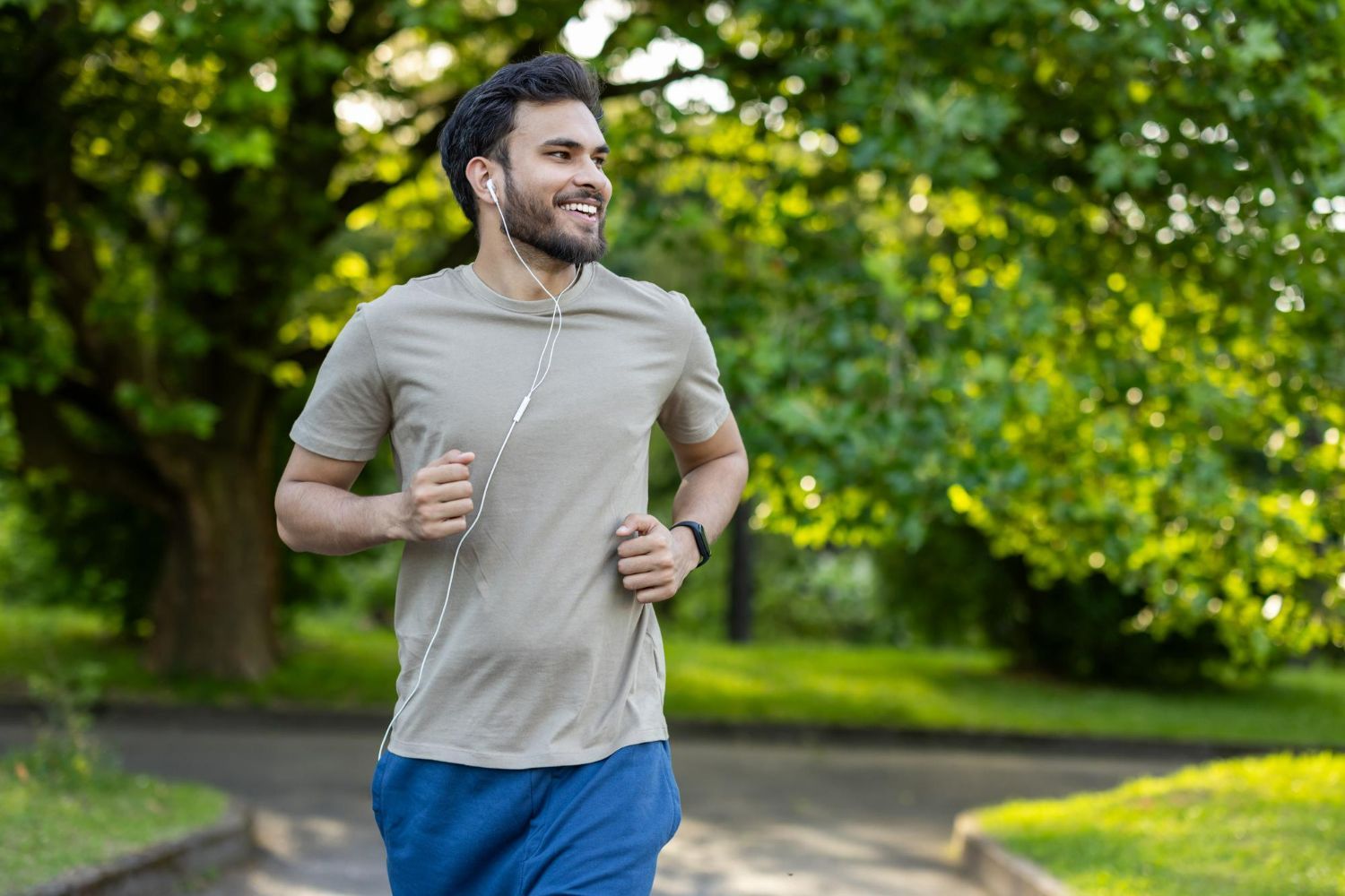 Man jogging outdoors, smiling, wearing earbuds, light-colored shirt, and blue shorts. Green trees in the background.
