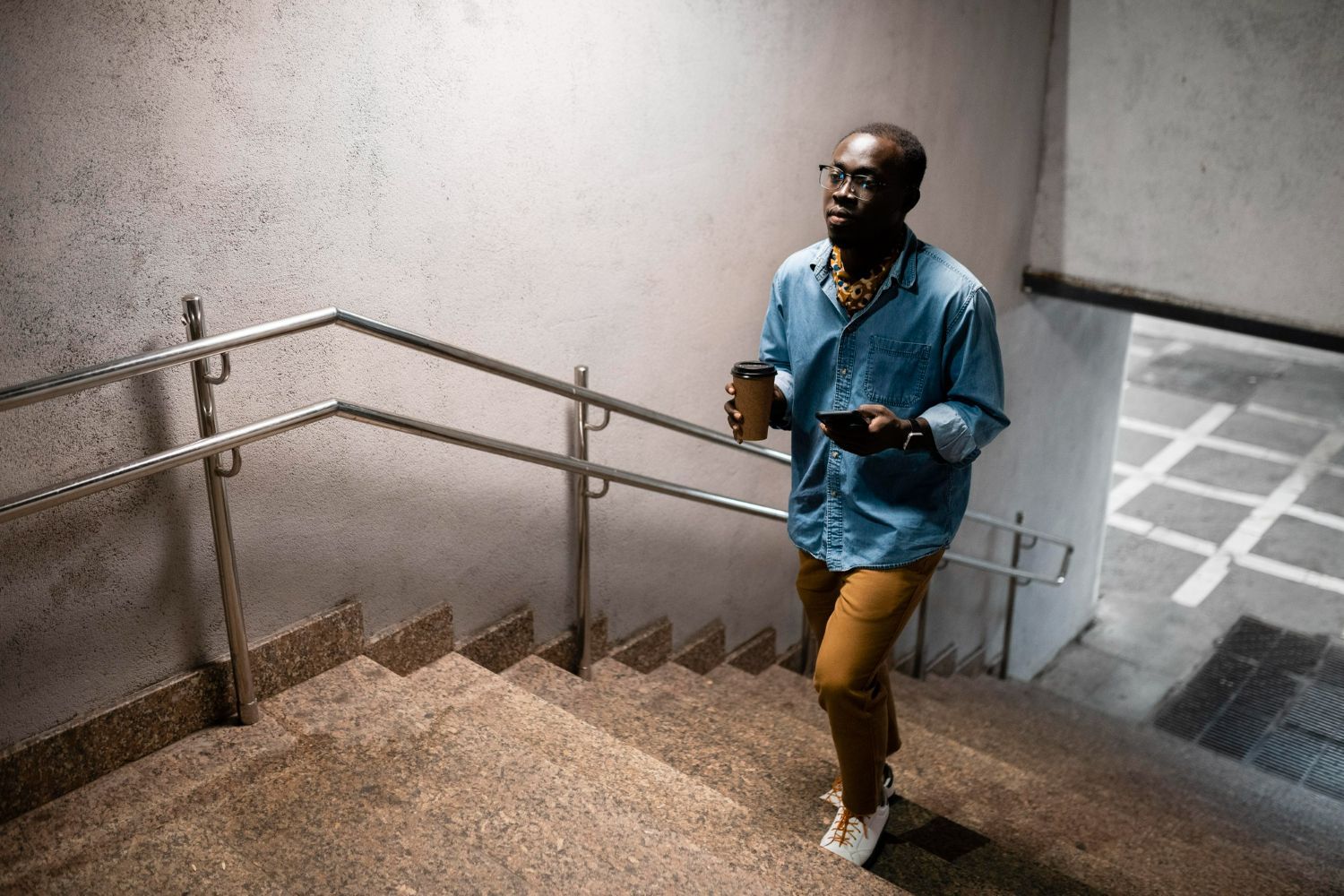 A Black man climbs stairs, holding coffee and a phone. He wears a blue shirt and tan pants, in a concrete setting.