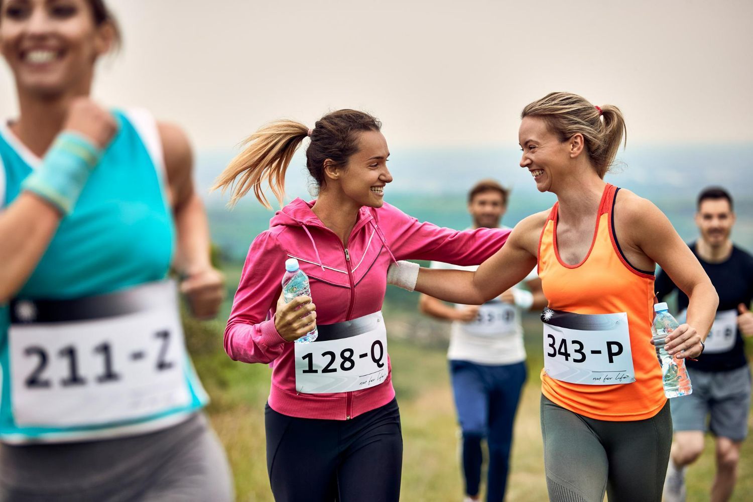 Runners in a race, two women smiling and touching each other, one holding a water bottle.
