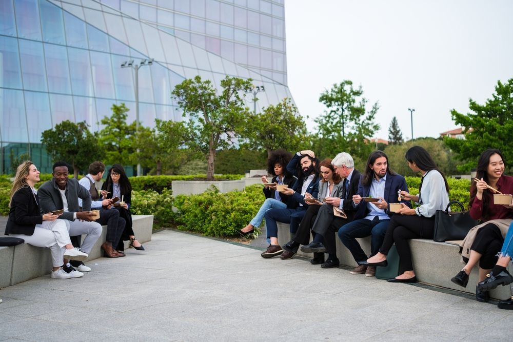 Group of diverse people seated outdoors, looking at their phones, in front of a modern building.