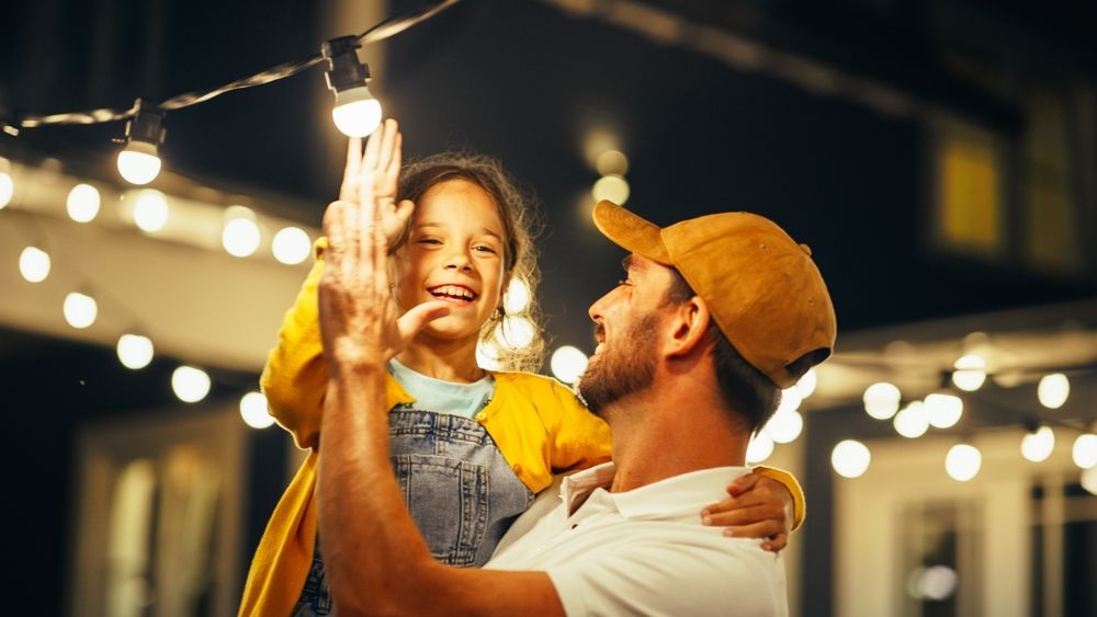 Father holds daughter high, giving her a high five under string lights at night. They are smiling.