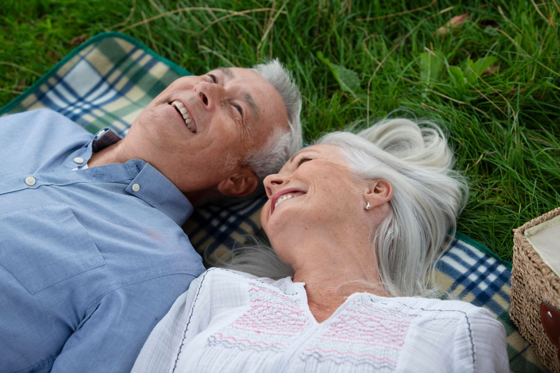 An elderly couple is laying on a blanket in the grass.