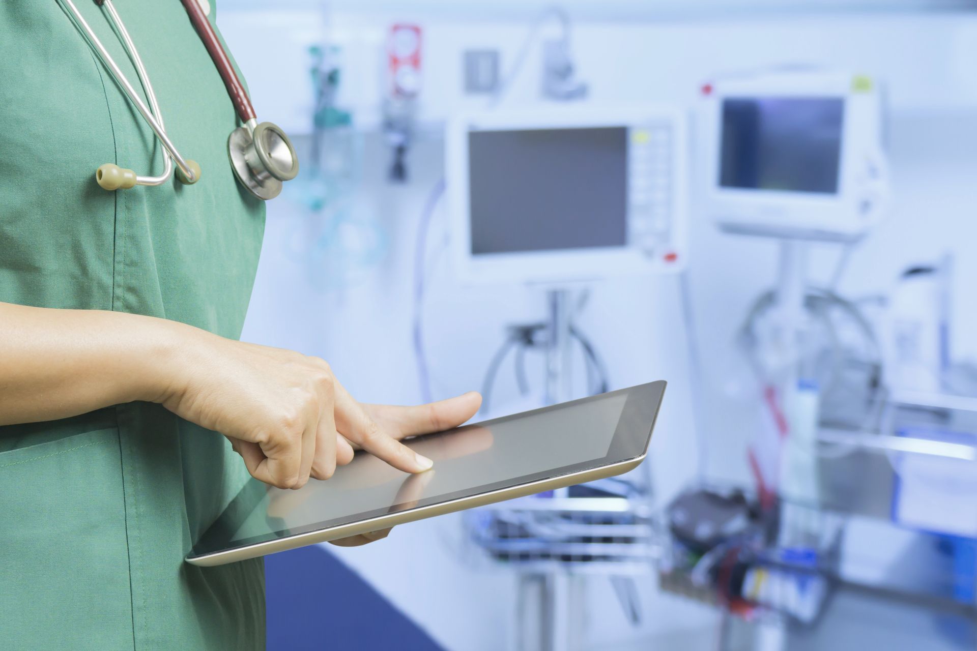 Persoon in groene operatiekleding gebruikt een tablet in een medische ruimte met apparatuur.