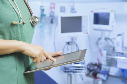 Persoon in groene operatiekleding gebruikt een tablet in een medische ruimte met apparatuur.