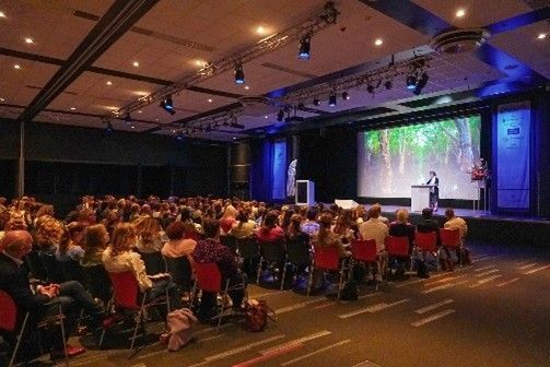 Het publiek op een conferentie luistert naar een spreker op het podium, waar een scherm een ​​afbeelding van een bos toont.