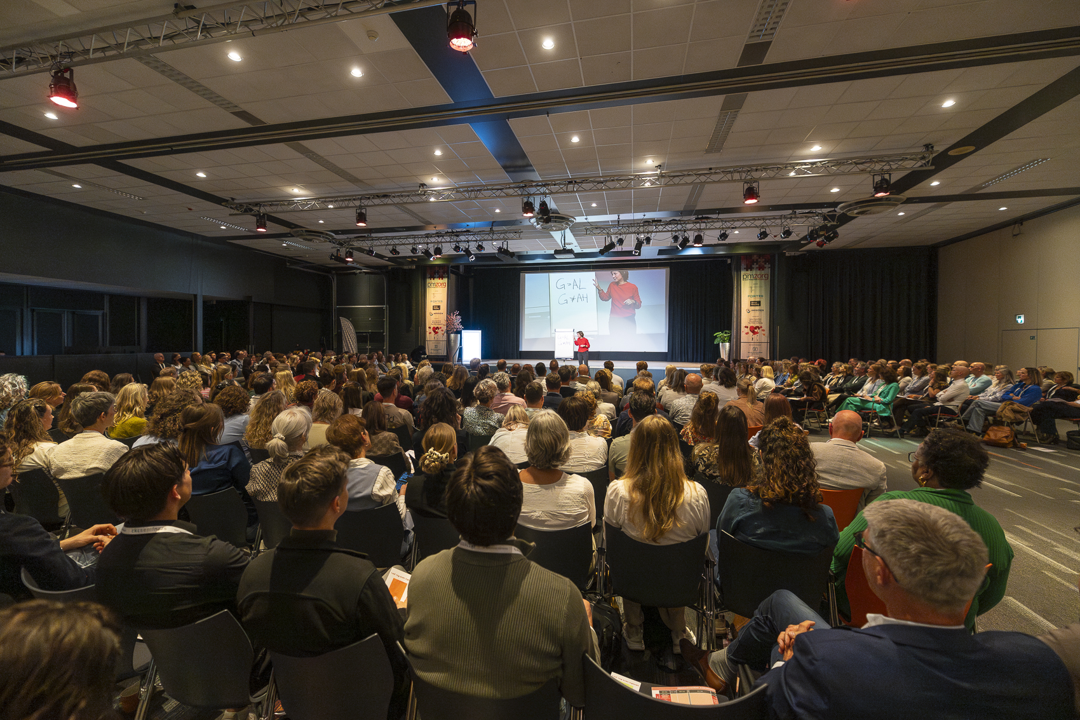 Het publiek op een conferentie luistert naar een spreker op het podium, waar een scherm een ​​afbeelding van een bos toont.