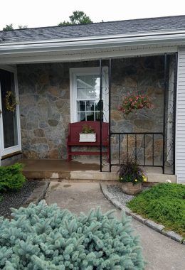 Stone-walled porch with red bench, black railing, and hanging flower basket. Concrete path and blue-green bush.