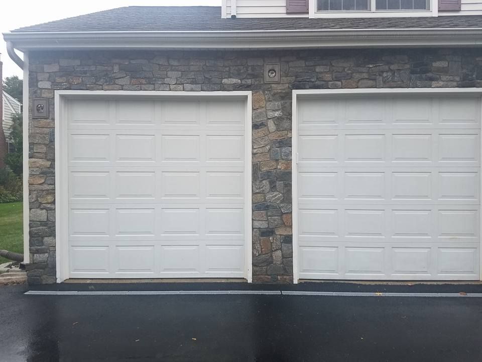 Two white garage doors on a stone-faced building, set on a dark asphalt surface.