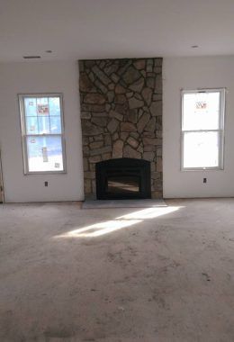 Stone fireplace centered in a room with two windows. Unfinished floor, white walls.