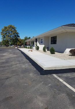 Concrete sidewalk leading to a light-colored building with dark windows, blue sky.