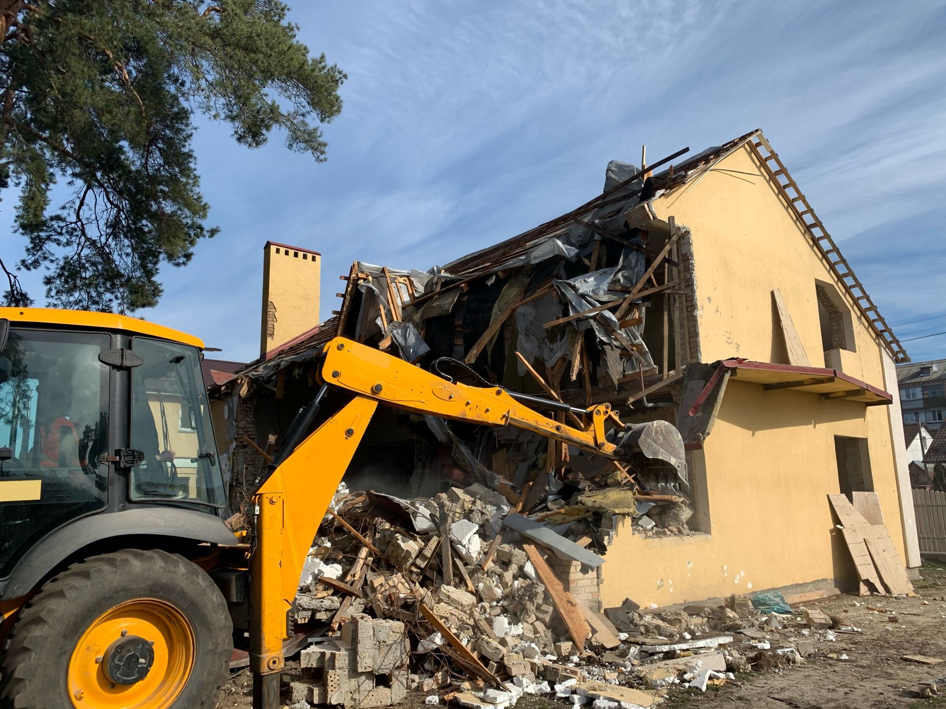 Yellow excavator demolishing a yellow building with its roof destroyed on a sunny day.