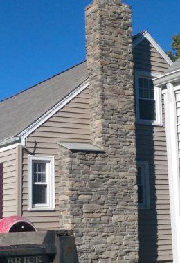 Stone chimney on a house with gray siding and windows, against a blue sky.