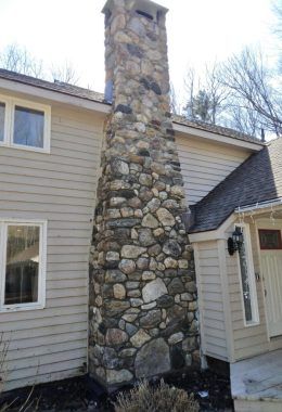 Stone chimney on a light-colored house. Brown roof and siding. Clear sky in background.