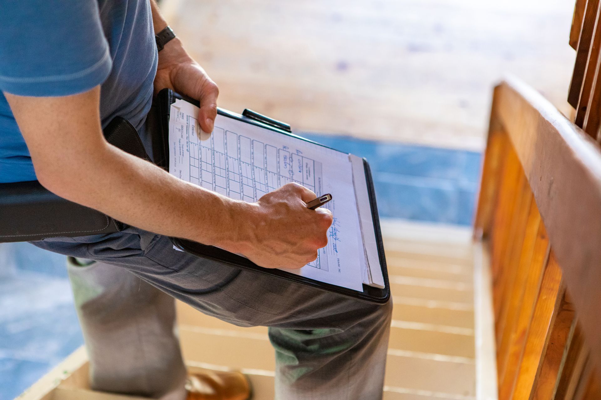 A close-up and high-angle view of a professional male wearing a blue t-shirt, diligently filling out inspection forms while standing on a flight of stairs during a home inspection.