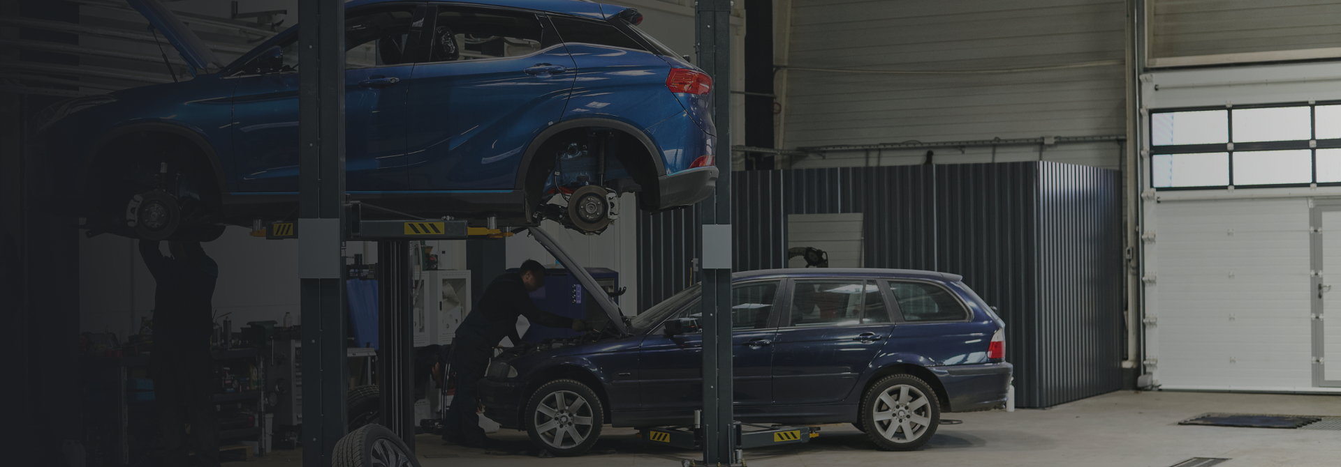 Blue car on a lift in a garage, a mechanic works on another car below. | Bama Direct Automotive Llc