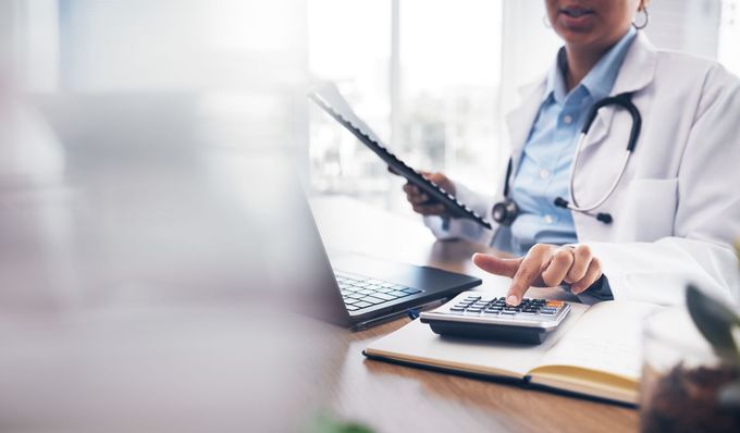 A doctor is sitting at a desk using a laptop and a calculator. - Newport News, VA - Precision Pro Coders