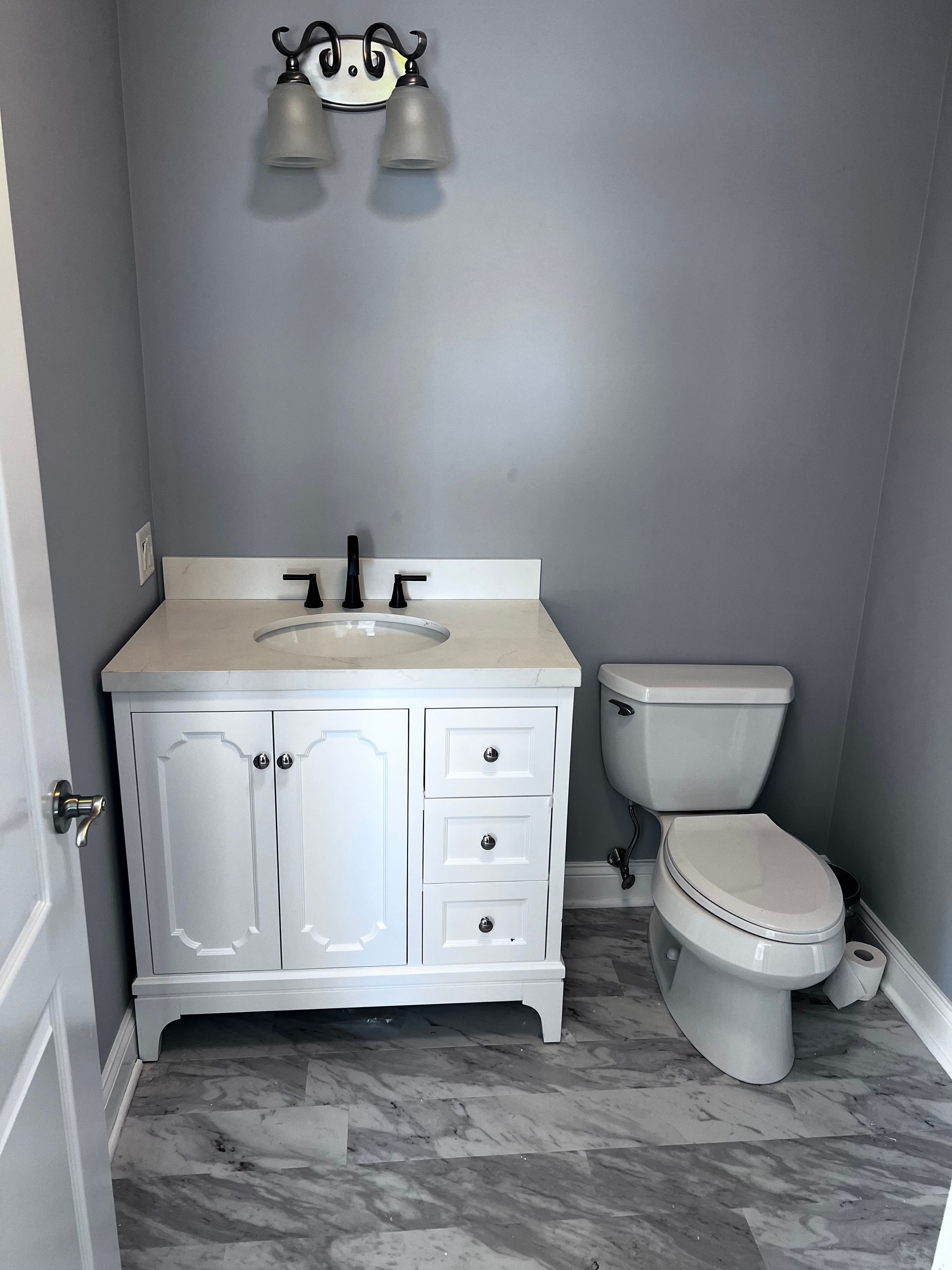 Small gray bathroom with white vanity, toilet, and wall light over a marble-look floor.