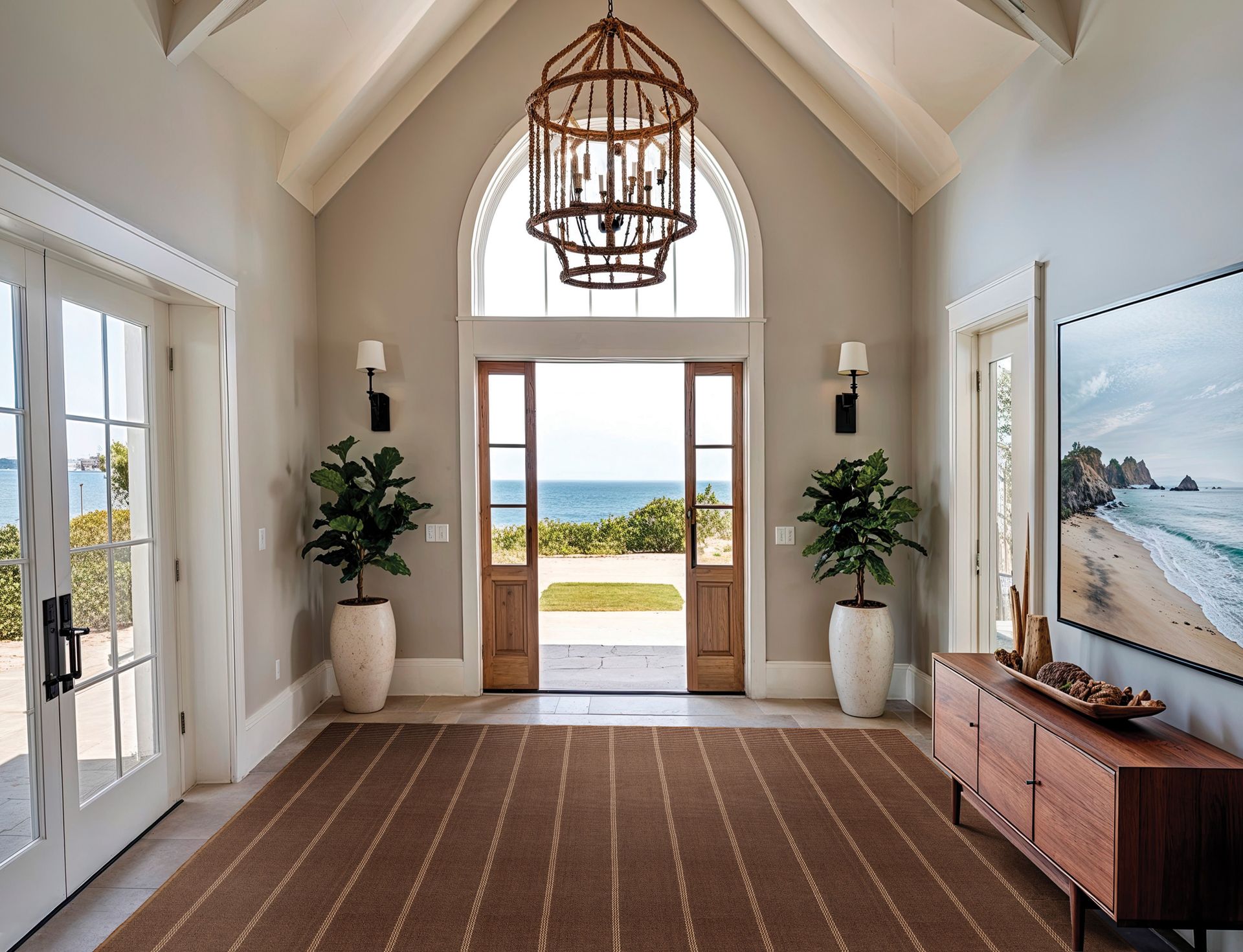 Elegant entryway with ocean view through open double doors, rug, potted plants, chandelier, and a wood console.