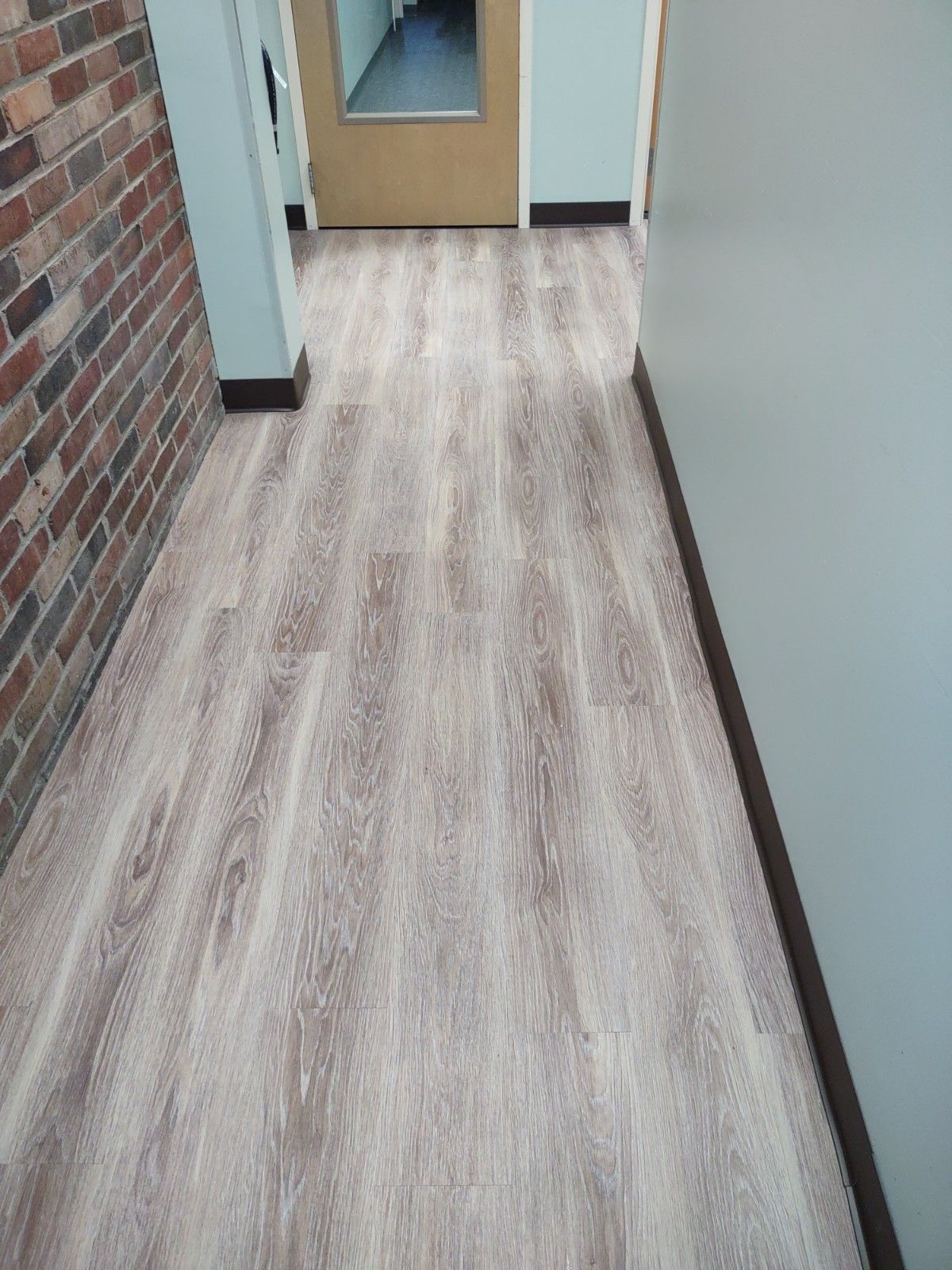 Empty hallway with wood-pattern flooring, brick wall on the left, and a door at the end.
