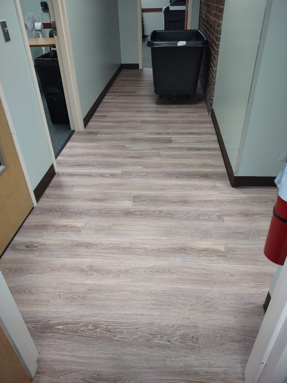 Office hallway with light wood flooring, cabinets along the sides, and a black trash bin at the end.