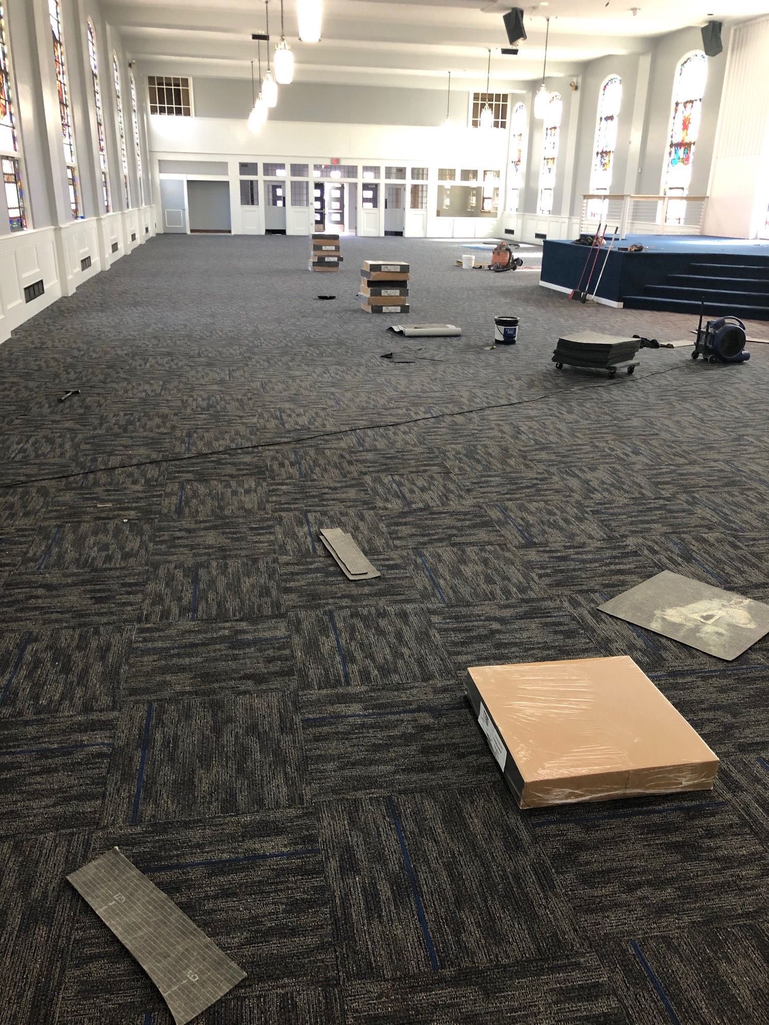 Empty hall with patterned carpet, scattered floor panels, and stacked chairs near large windows.