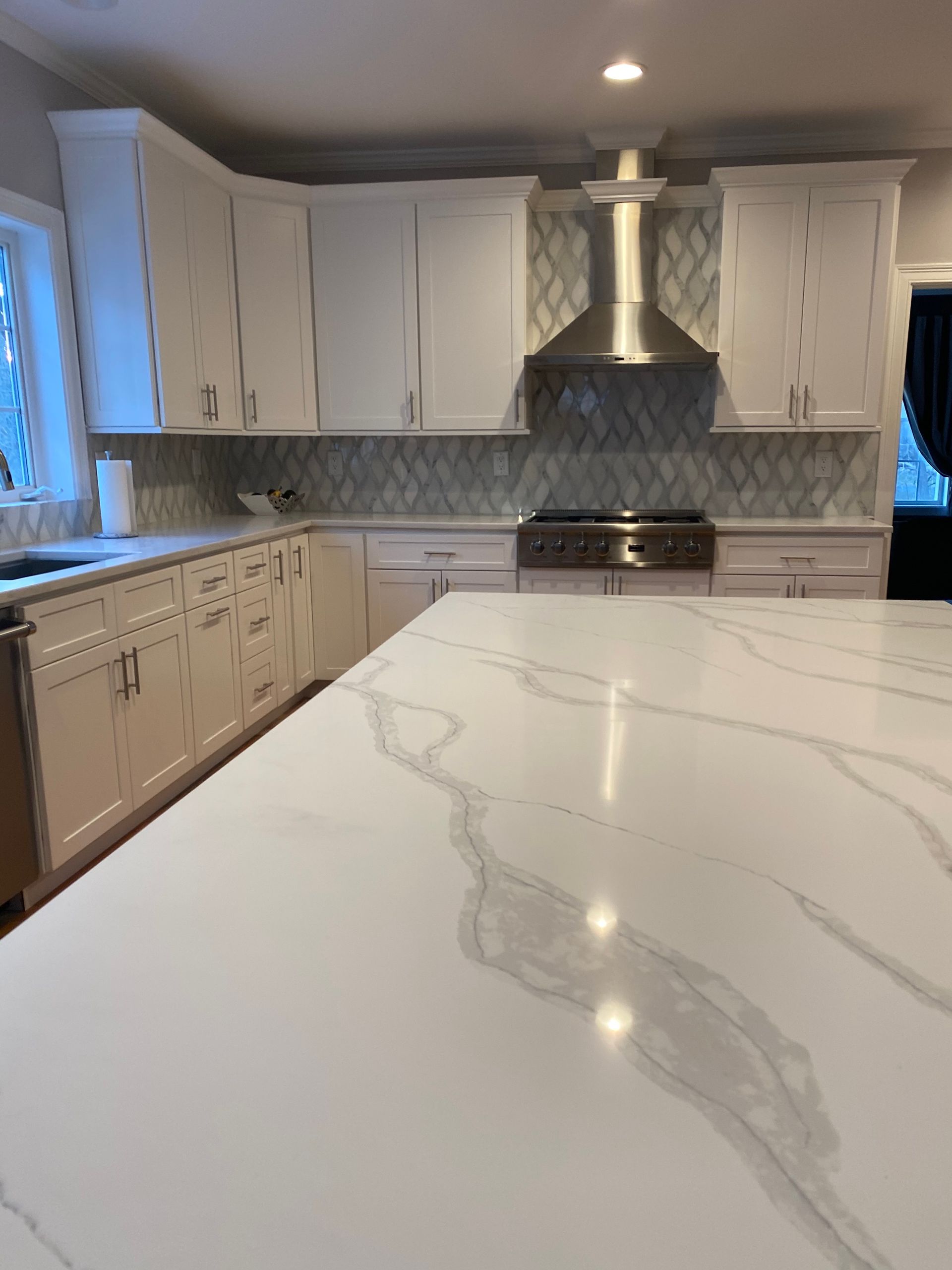 Bright white kitchen with marble island, light cabinets, stainless range hood, and tiled backsplash.