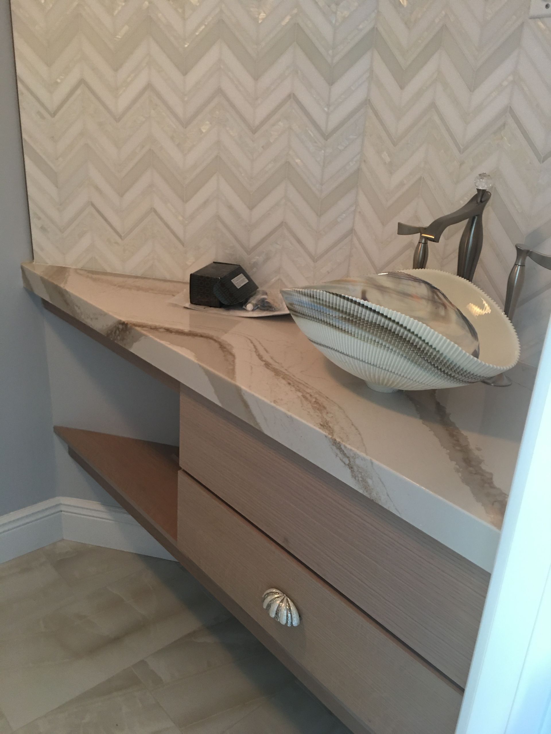 Bathroom vanity with beige marble countertop, sink, faucet, and drawer storage against herringbone wallpaper