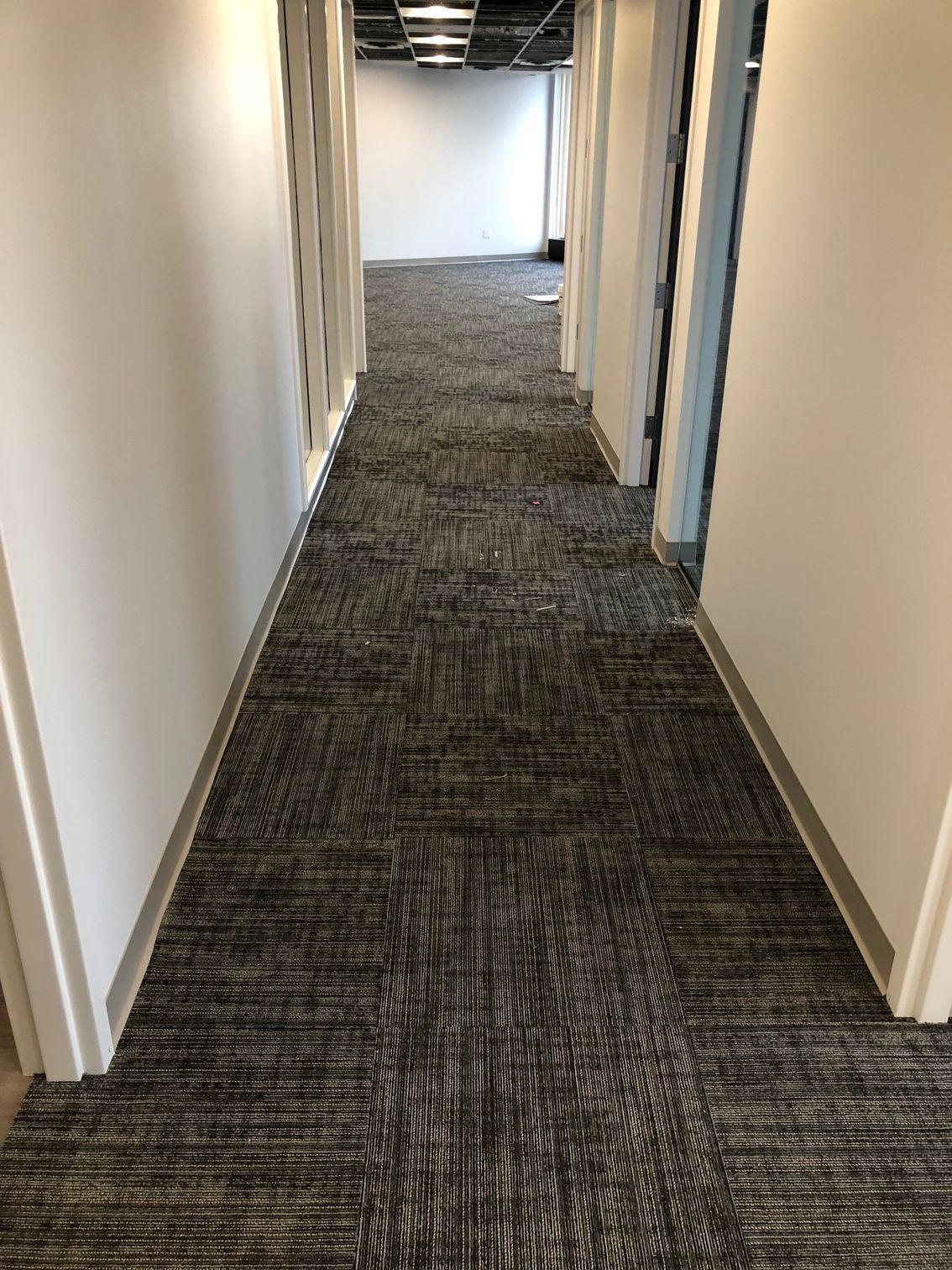 Narrow office hallway with gray carpet and white walls, lit by overhead lights.