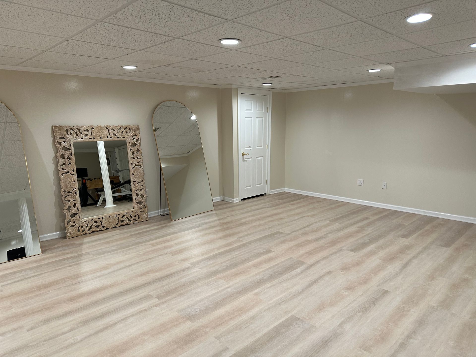 Empty beige room with light wood flooring, mirrored wall accents, and a white door.