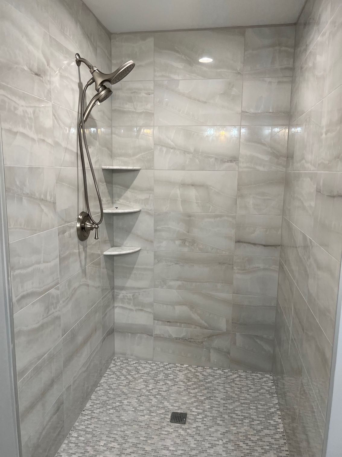 Modern tiled walk-in shower with handheld showerhead, corner shelves, and pebble-style floor.