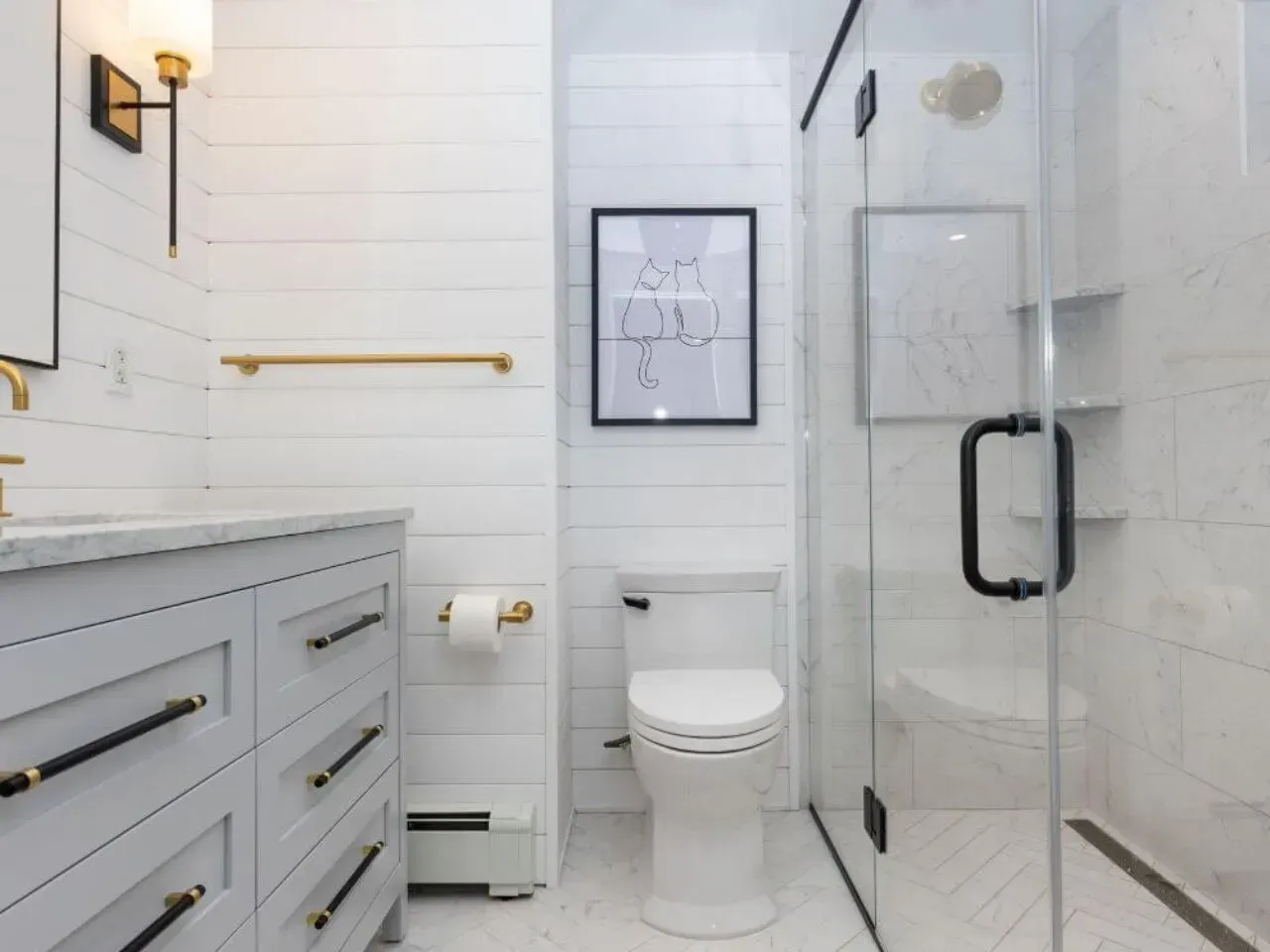 A small, modern white bathroom with gold and black accents. Includes a vanity, toilet, and glass shower.