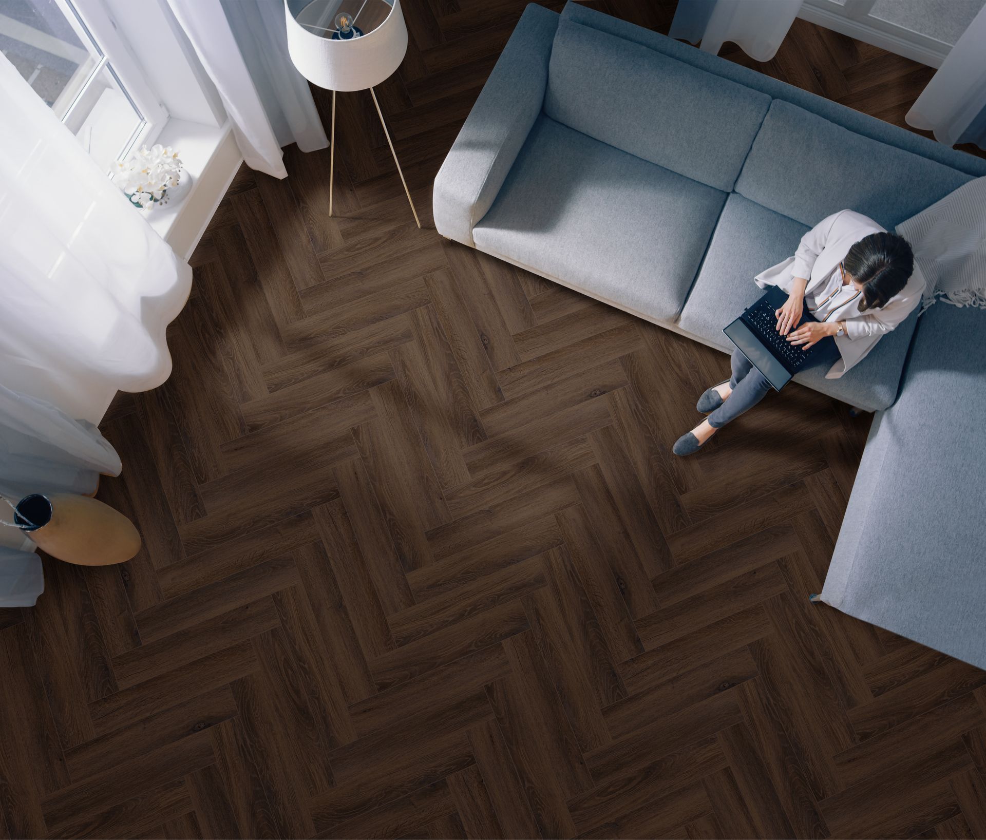 Person sitting on a gray sofa in a sunlit living room with a dark herringbone wood floor.