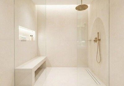 Modern, cream-colored bathroom with a walk-in shower. Features a bench, rain showerhead, and gold fixtures.