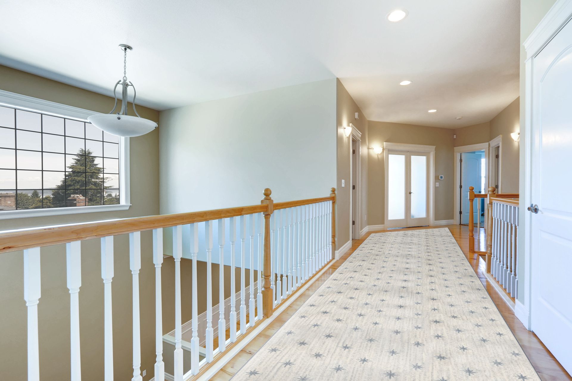 Hallway with a cream-colored runner rug and white banister. Large window on left, several doors on right.