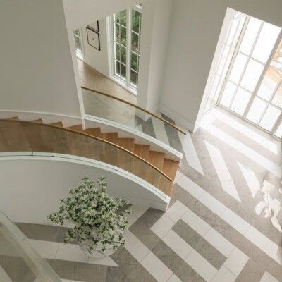 High-angle view of a bright foyer with a curved wooden staircase and geometric patterned floor. Large windows provide natural light.