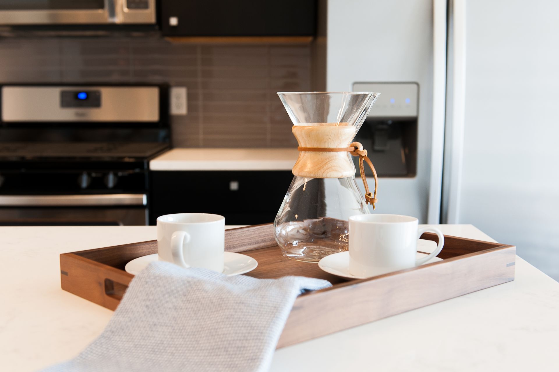 A wooden tray with two cups of coffee and a coffee maker on a kitchen counter.