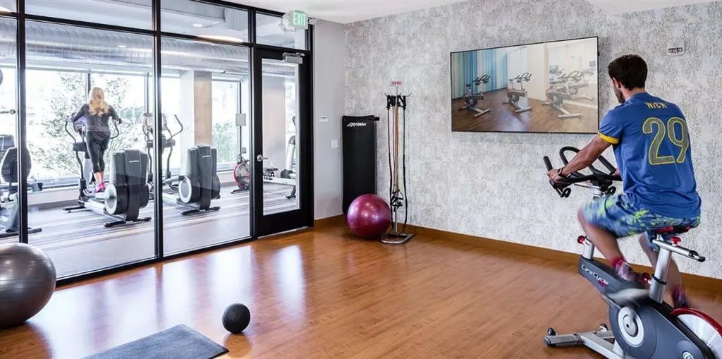 A person on a stationary bike in a gym with other exercise equipment and someone else on an elliptical machine.